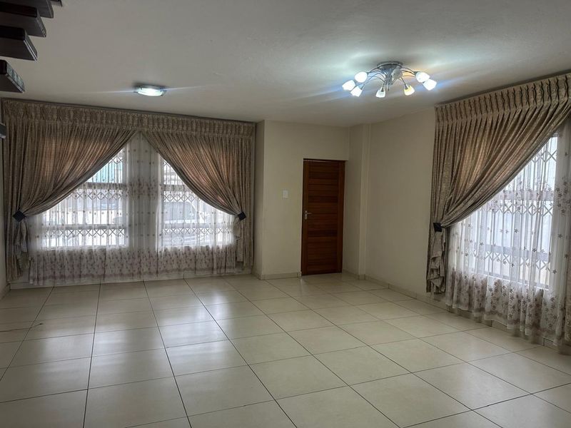 A spacious, empty living room featuring neutral tiled flooring, large windows with security bars, and dual ceiling light fixtures.