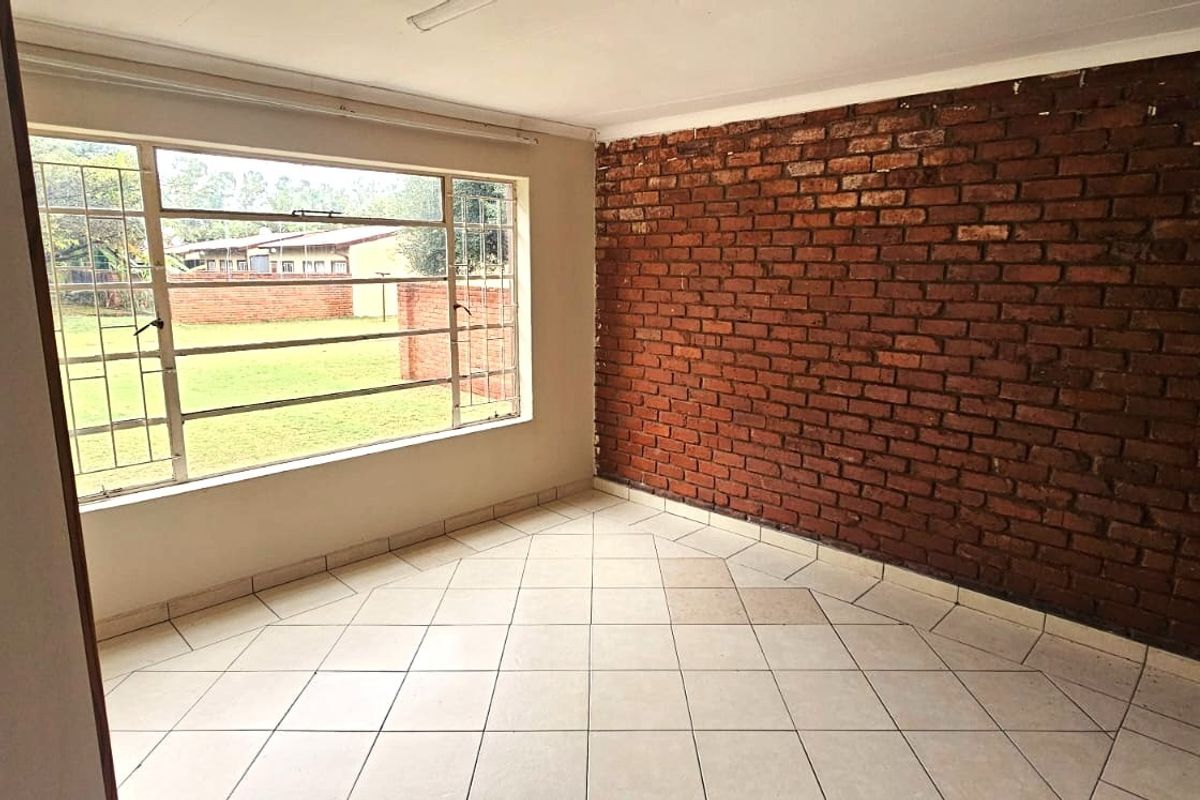 An empty living room featuring a rustic exposed brick wall, tiled flooring, and a large window providing natural light and garden views.
