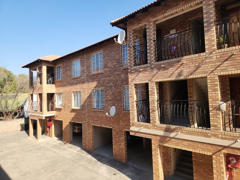 Exterior view of a multi-story face brick apartment building featuring covered parking bays, private balconies with wrought iron railings, and a tiled roof.