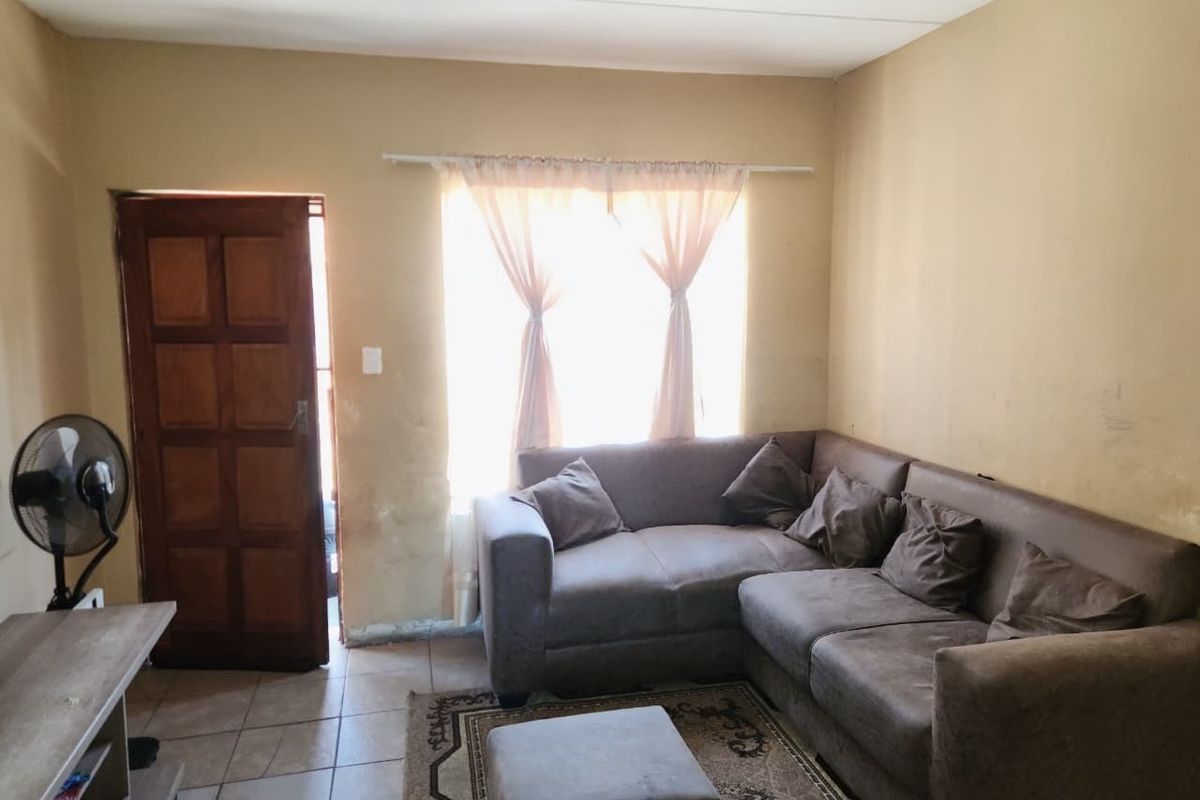 A living room featuring tiled flooring, a wooden entrance door, and a window, currently showing signs of wear on the walls and finishes.