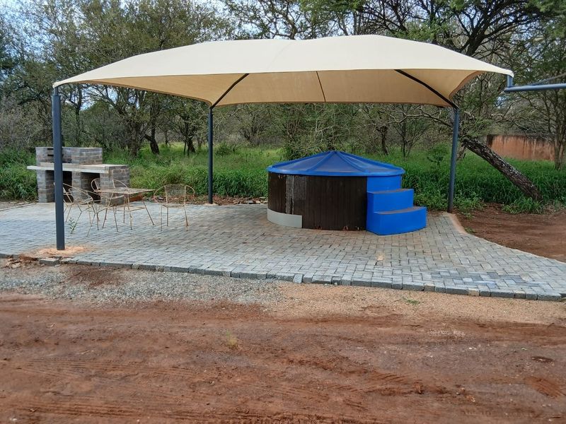 Outdoor entertainment area featuring a paved patio, shade sail, built-in braai, and a small plunge pool set in a natural farm landscape.