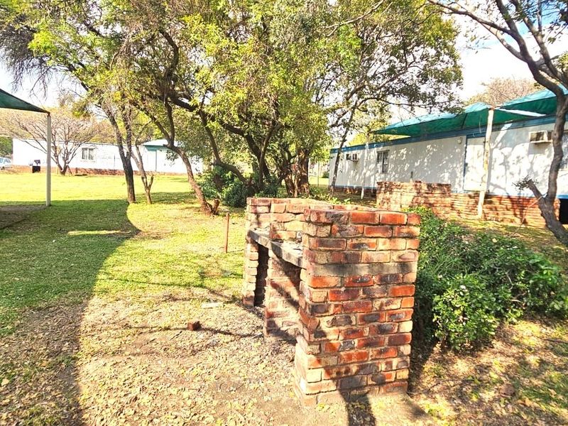 Spacious outdoor garden area featuring a built-in brick braai, mature trees, and multiple prefabricated structures with shade cloth coverings.