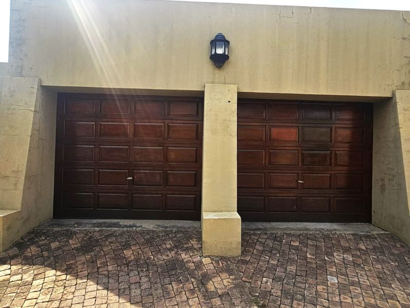 A double garage with wooden doors and a paved driveway, featuring exterior wall lighting and visible plaster cracking on the facade.