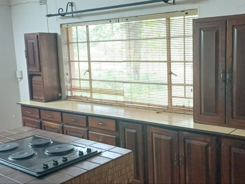 A dated kitchen featuring wooden cabinetry, a tiled island with a built-in electric hob, and a large window with venetian blinds.