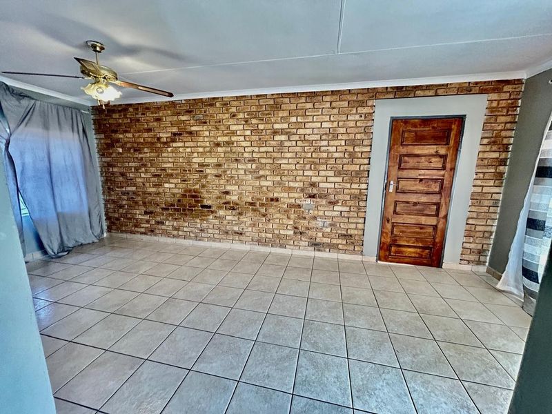 A spacious living area featuring a prominent exposed brick feature wall, tiled flooring, and a ceiling fan.