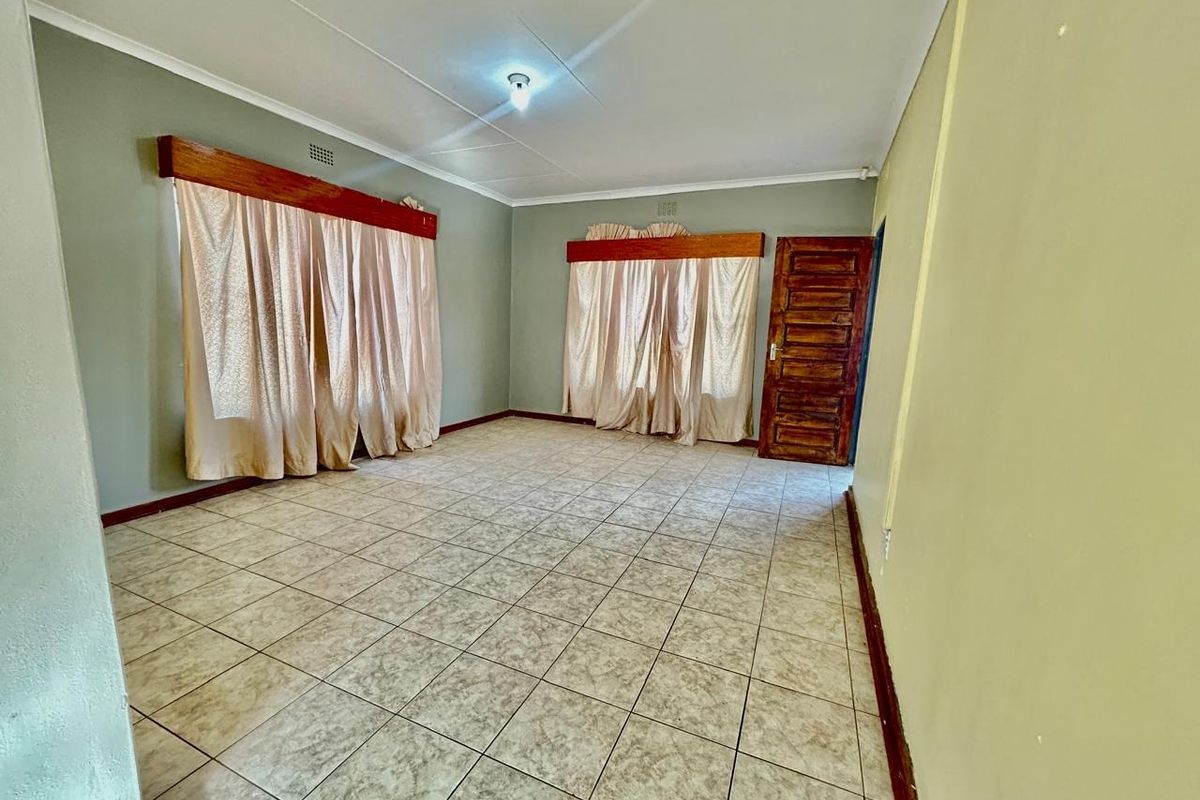 A spacious, empty living room featuring neutral tiled flooring, wooden door, and classic wooden curtain pelmets, offering a blank canvas for interior design.