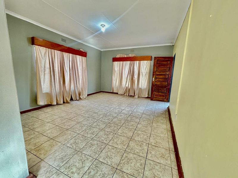 A spacious, empty living room featuring neutral tiled flooring, wooden door, and classic wooden curtain pelmets, offering a blank canvas for interior design.