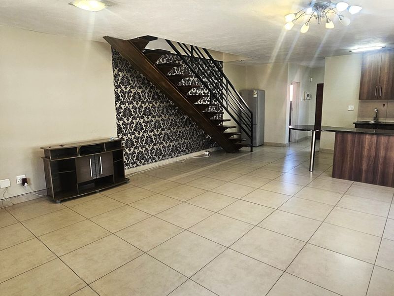 Open-plan living area featuring tiled floors, a central staircase with metal railing, and a functional kitchen with wood-finish cabinetry and an island.