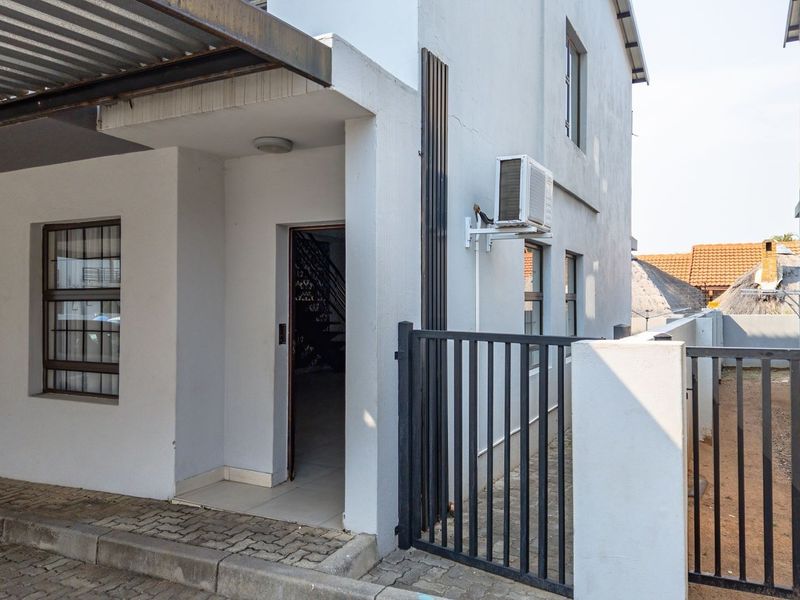 Front exterior view of the townhouse featuring a covered carport, paved driveway, security gate, and wall-mounted air conditioning unit.