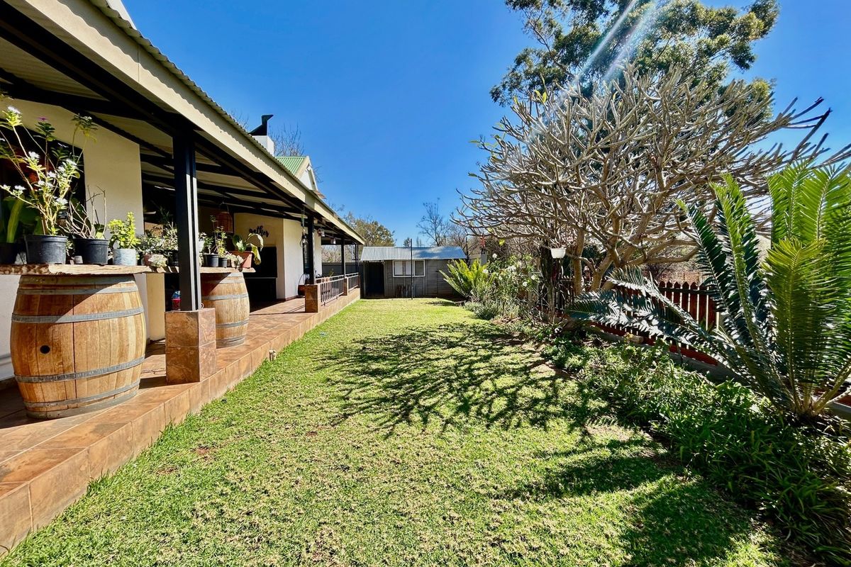 A spacious rear garden featuring a covered patio with tiled flooring, a built-in braai area, and a detached outbuilding at the rear.