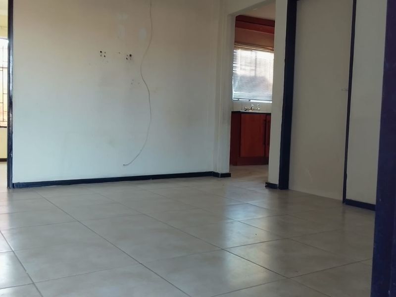 An empty living area featuring tiled flooring and visible wall damage, with a view through to an adjacent kitchen space.