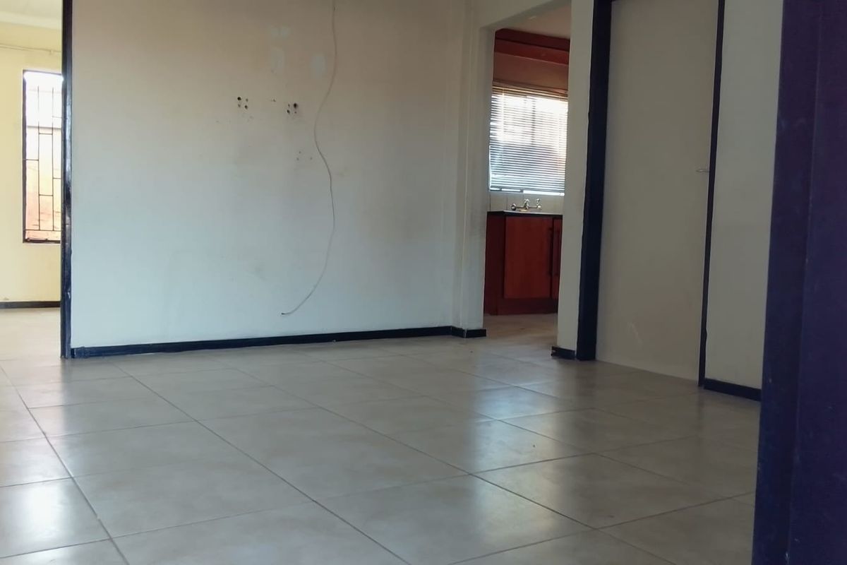 An empty living area featuring tiled flooring and visible wall damage, with a view through to an adjacent kitchen space.