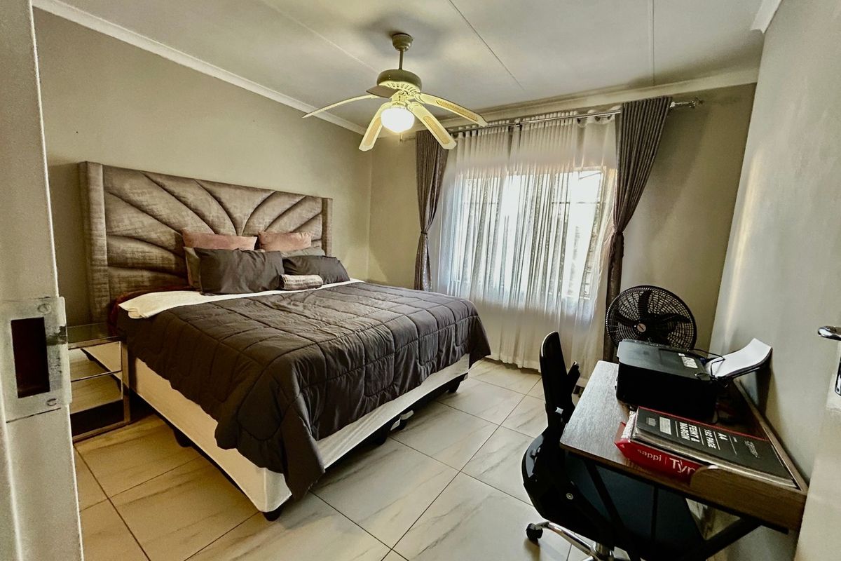 A well-maintained bedroom featuring neutral tiled flooring, a central ceiling fan, and large windows with fitted curtain rails.