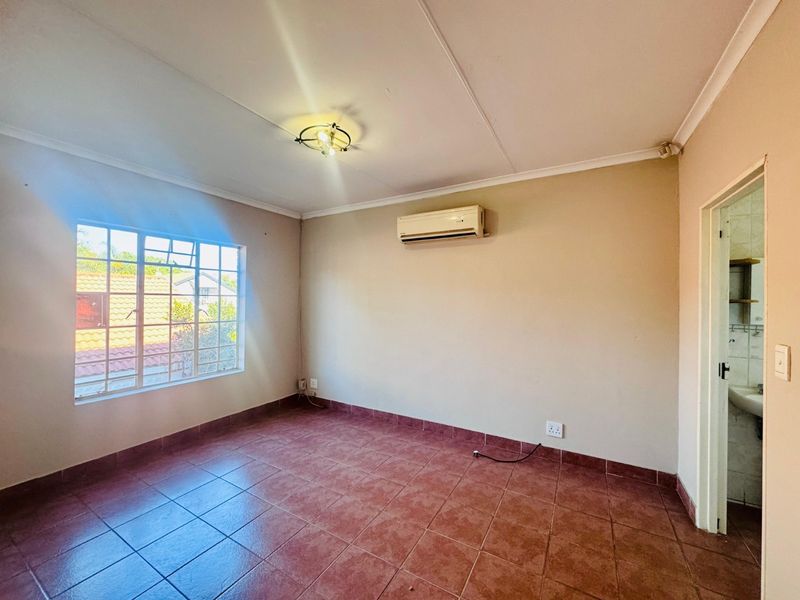 An empty bedroom featuring tiled flooring, a wall-mounted air conditioning unit, and a grid-pattern window, with an adjacent bathroom visible through the doorway.