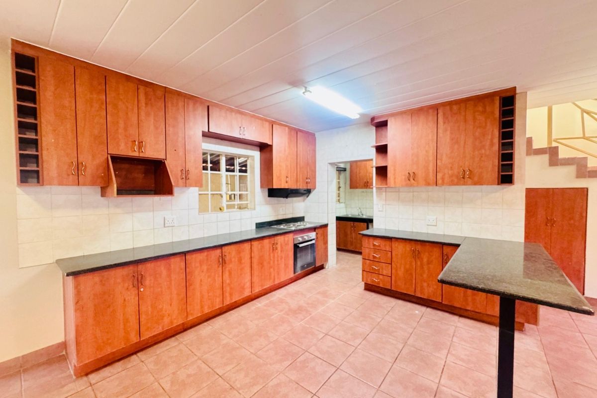 Spacious kitchen featuring extensive wooden cabinetry, dark countertops, built-in oven and hob, tiled flooring, and a breakfast bar, with an adjacent scullery area.