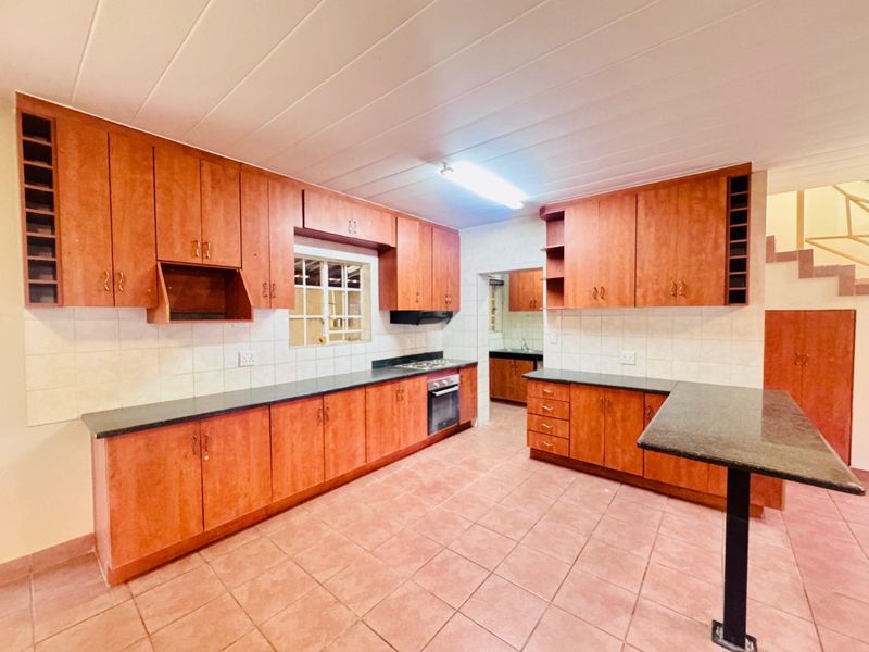 Spacious kitchen featuring extensive wooden cabinetry, dark countertops, built-in oven and hob, tiled flooring, and a breakfast bar, with an adjacent scullery area.