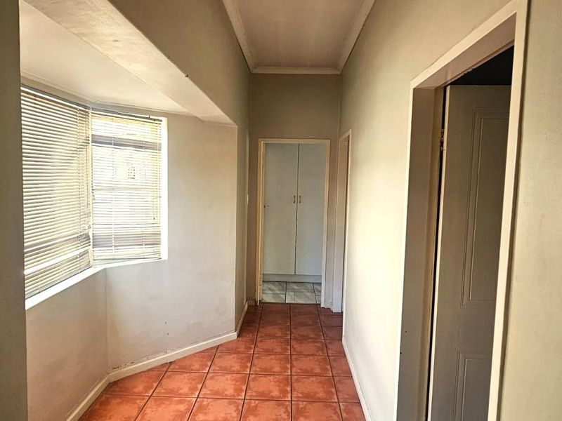 A vacant hallway featuring tiled flooring, a bay window with fitted blinds, and built-in storage cupboards, showing signs of wear.