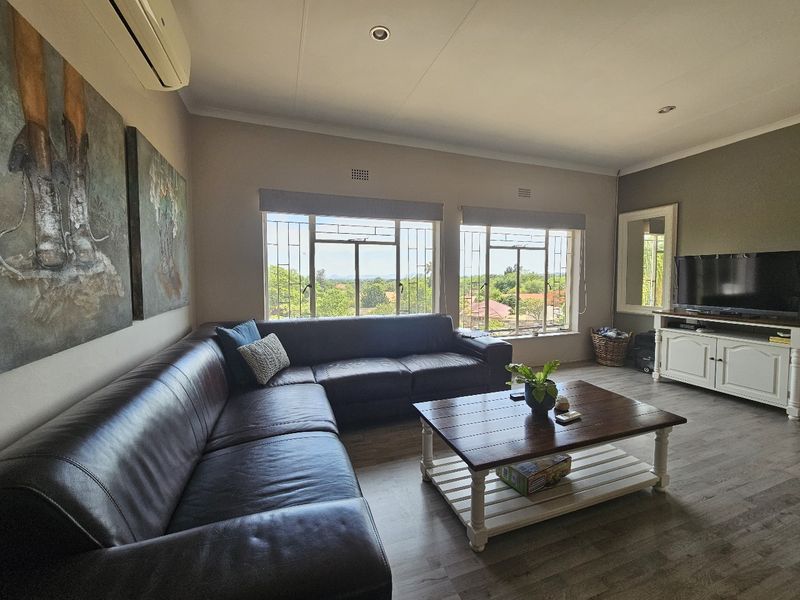 A comfortable living room featuring laminate flooring, recessed downlighting, a wall-mounted air conditioner, and large windows with security bars offering pleasant views.