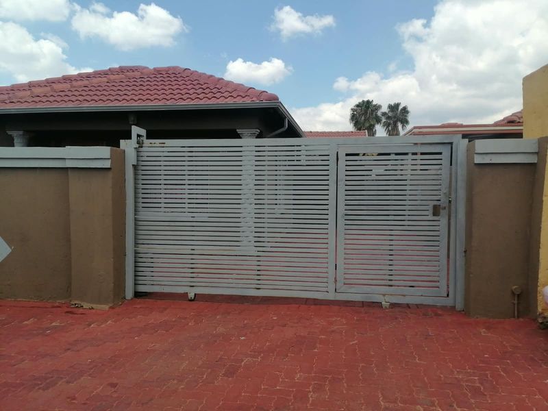 Front entrance view featuring a grey metal sliding gate, pedestrian gate, and brick boundary wall with a paved driveway.