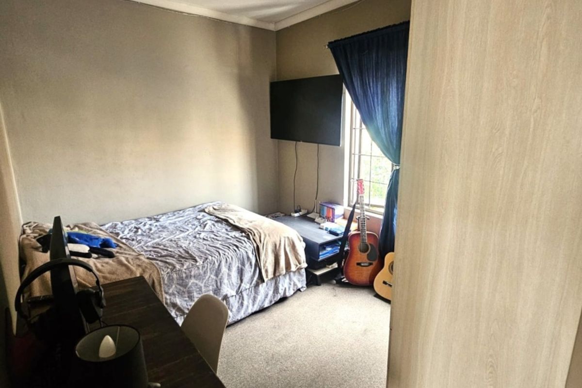 A compact bedroom featuring carpeted flooring, a built-in cupboard, and a window with curtaining, offering a functional space in need of cosmetic updates.