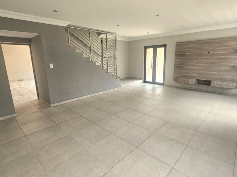Spacious open-plan living area featuring neutral tiled flooring, recessed lighting, a modern built-in media unit, and a staircase with a sleek stainless steel balustrade.