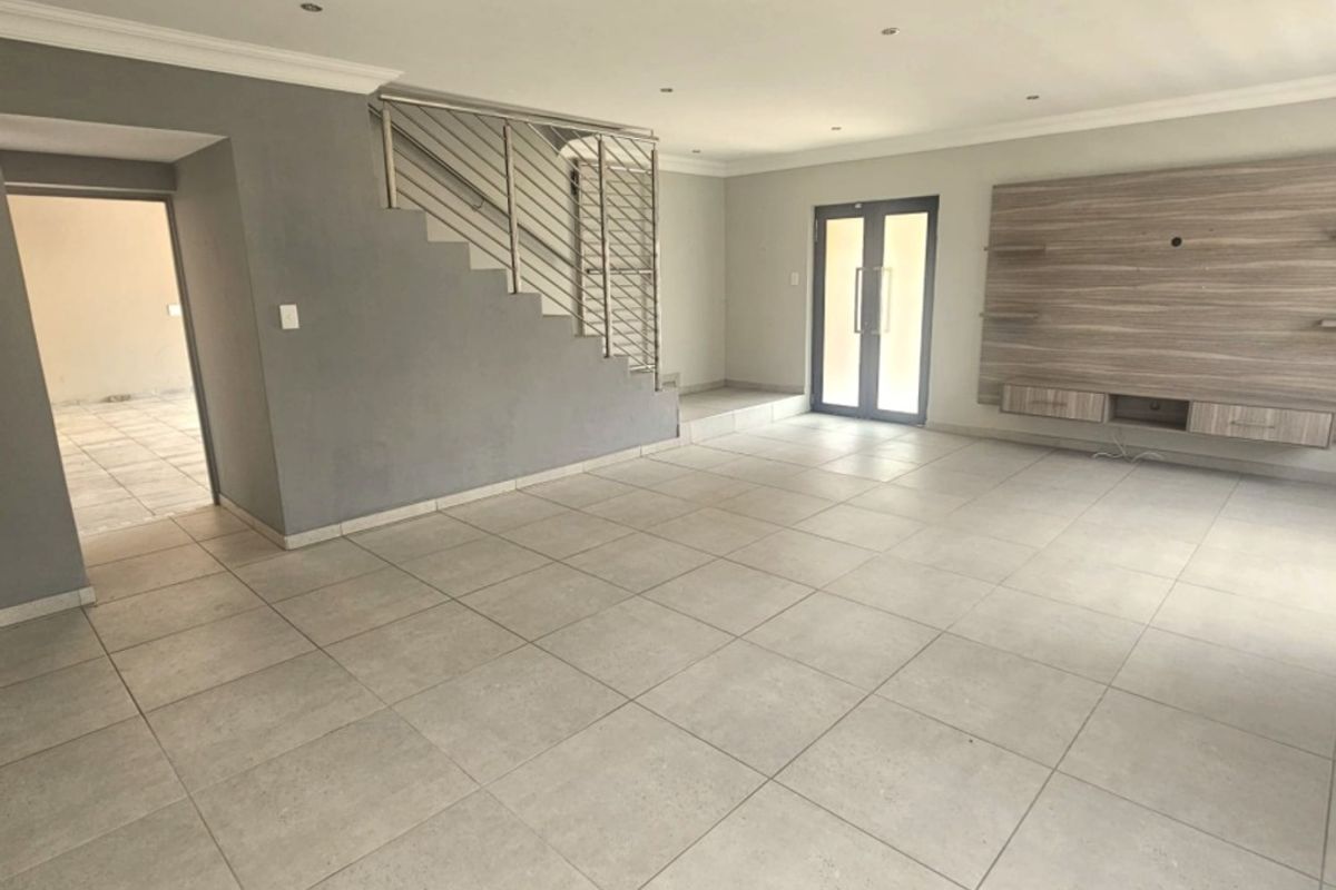 Spacious open-plan living area featuring neutral tiled flooring, recessed lighting, a modern built-in media unit, and a staircase with a sleek stainless steel balustrade.