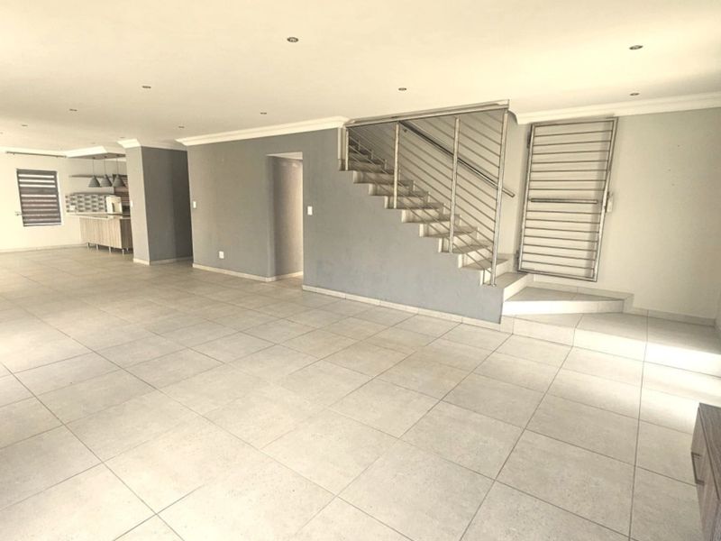 Spacious open-plan living area featuring neutral tiled flooring, recessed lighting, and a modern staircase with a stainless steel balustrade, leading to a kitchen area.