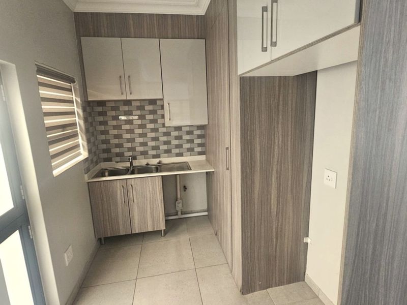 A clean and functional scullery featuring modern wood-grain cabinetry, a stainless steel sink, tiled backsplash, and neutral floor tiles.