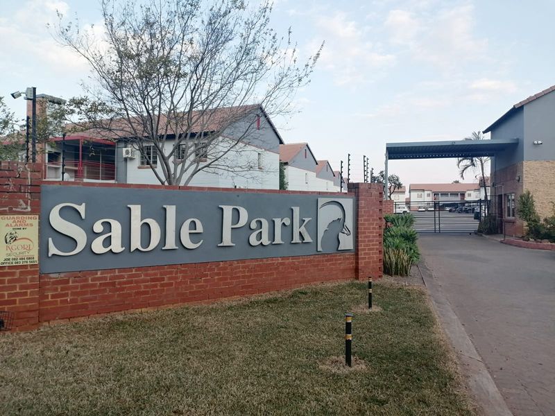 Entrance view of Sable Park complex featuring a brick boundary wall, security gate, and paved driveway access.