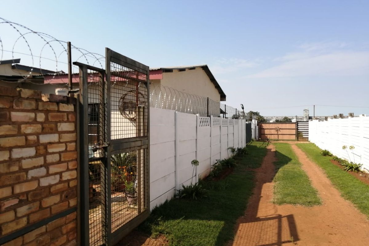 A side view of the property featuring a secure gated entrance, precast concrete boundary walling, and a dirt driveway leading to the rear.