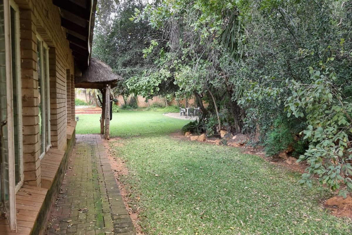 A view of the rear garden featuring a brick-paved walkway, mature trees, and a thatched-roof outdoor structure.