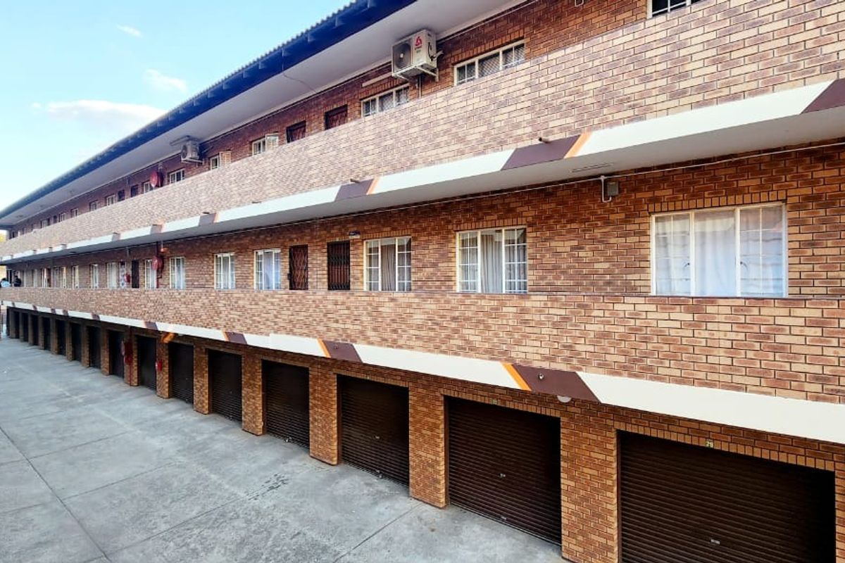 Exterior view of a multi-story apartment complex featuring face-brick facade, ground-level garage units, and upper-level balconies with integrated air conditioning units.