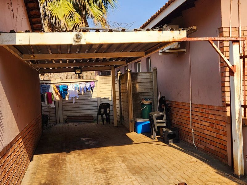 A paved outdoor carport area featuring a metal roof structure, brick boundary walls, and perimeter security fencing.