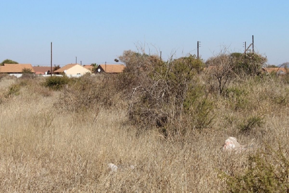 A view of an undeveloped residential plot featuring dry native vegetation with established residential housing visible in the background.