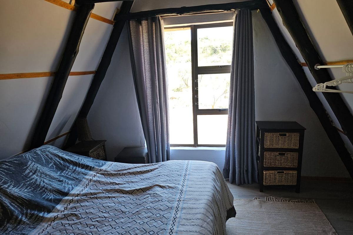 A cozy attic-style bedroom featuring exposed timber A-frame beams, a large window with natural light, and neutral wall finishes.