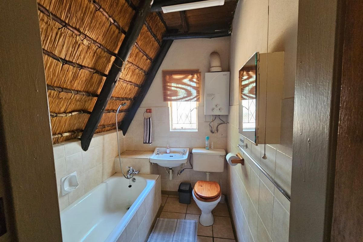 A compact bathroom featuring a bathtub, wall-mounted basin, toilet, and gas geyser, set under a traditional thatched roof with exposed timber beams.