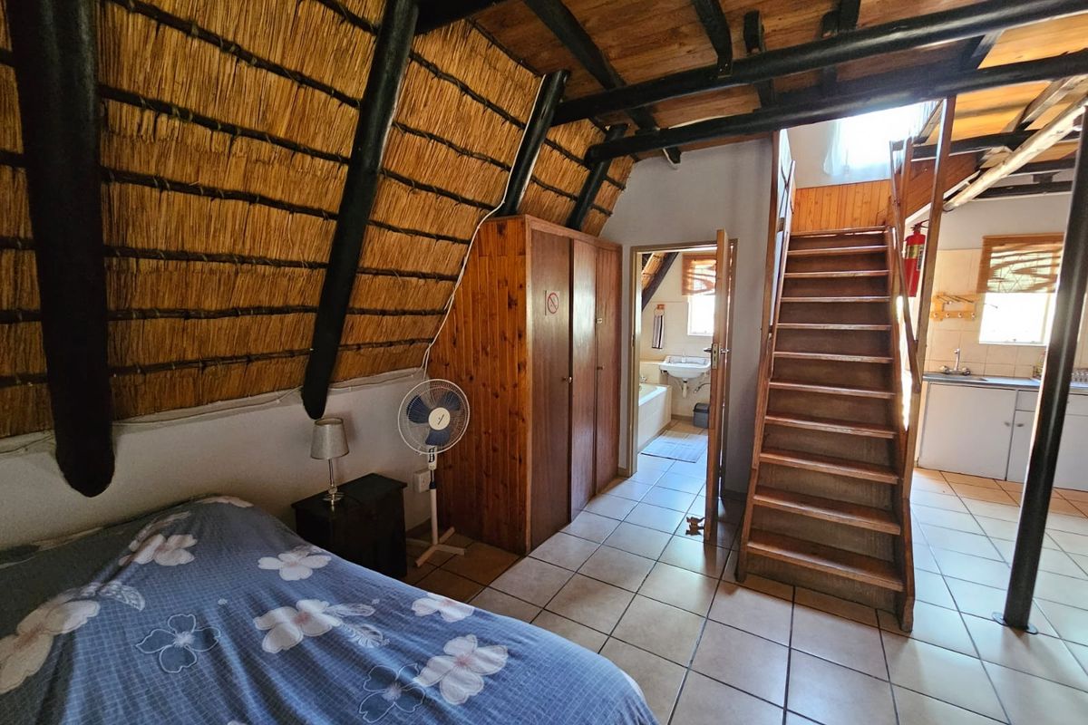 A bedroom featuring a thatched roof, exposed timber beams, tiled flooring, and an integrated staircase leading to an upper level with an adjacent bathroom.