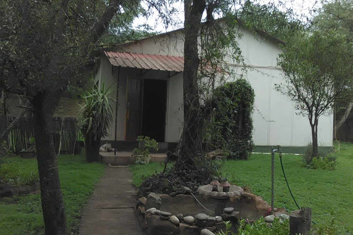 A view of a rural farm cottage exterior featuring a concrete pathway, established garden, and a small stone-bordered water feature.