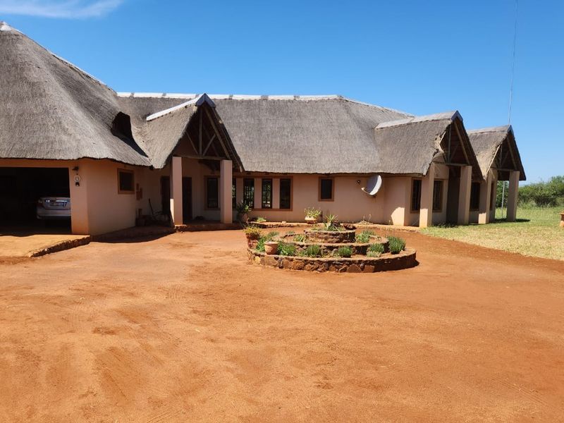 A spacious farm-style residence featuring a traditional thatched roof, covered entrance, and a large circular stone garden bed set within a wide dirt driveway.