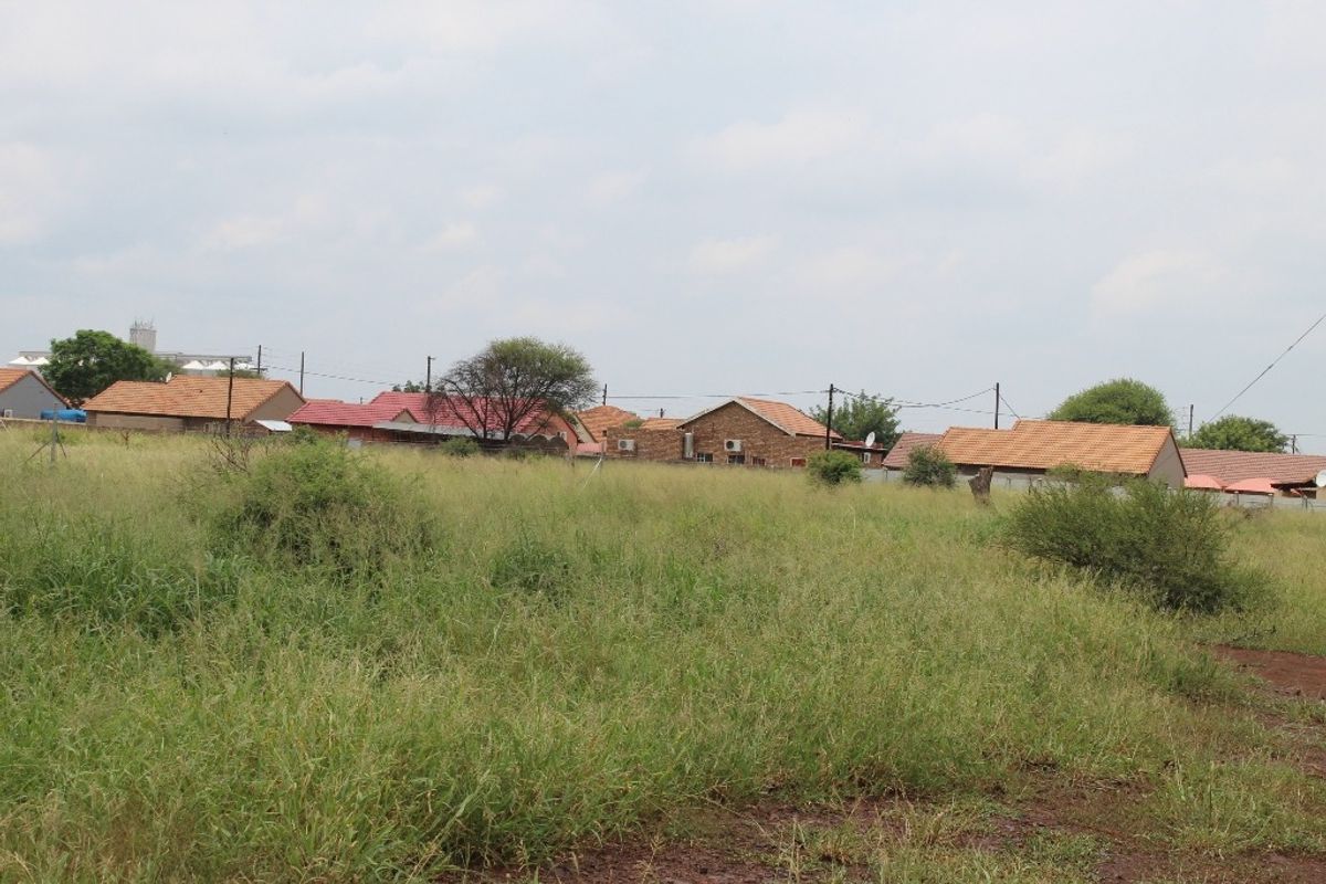 A view of a vacant residential plot featuring natural grass cover, with surrounding established residential properties visible in the background.