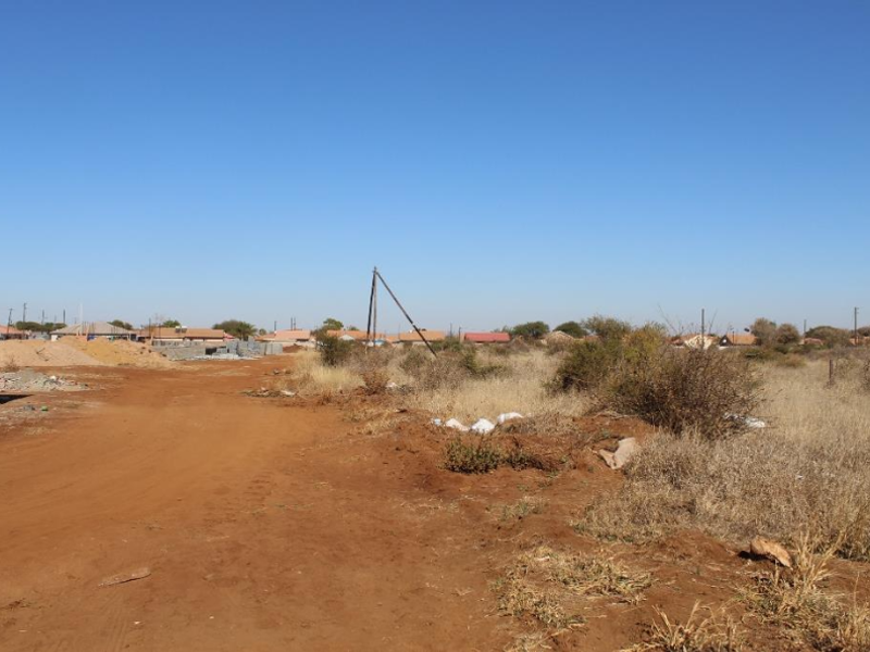 A view of an undeveloped residential plot featuring a dirt road, natural vegetation, and utility infrastructure in a suburban setting.