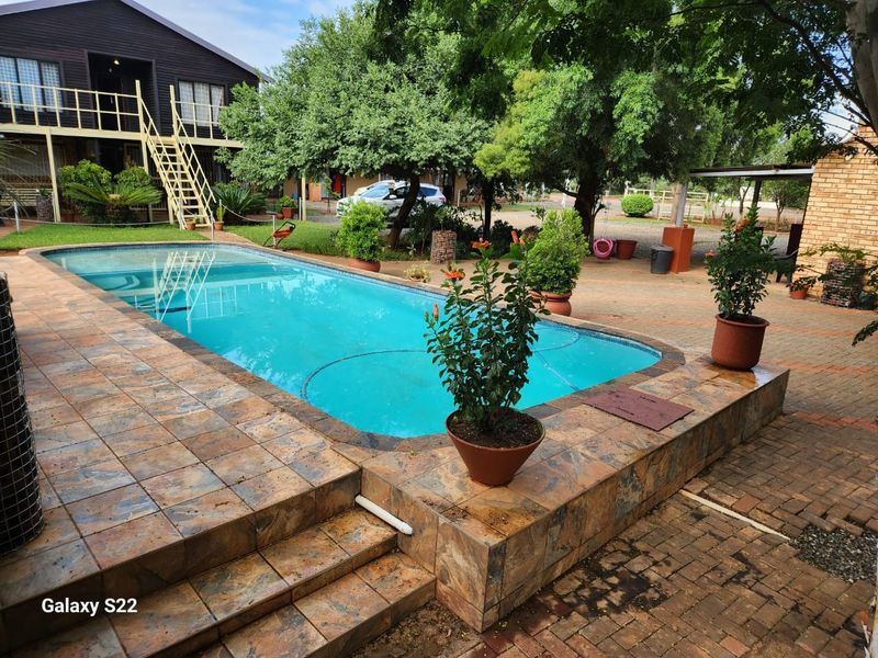 Outdoor pool area featuring a raised tiled pool deck, brick paving, and surrounding garden space with a multi-story building in the background.