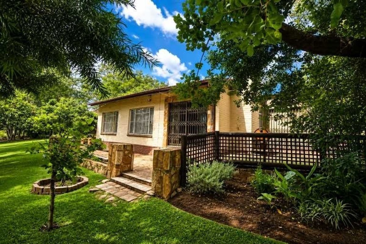 A well-maintained rear exterior view featuring a brick facade, paved patio area, stone pillars, and established garden surroundings with security features.