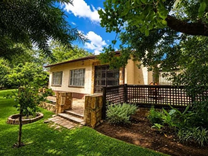 A well-maintained rear exterior view featuring a brick facade, paved patio area, stone pillars, and established garden surroundings with security features.