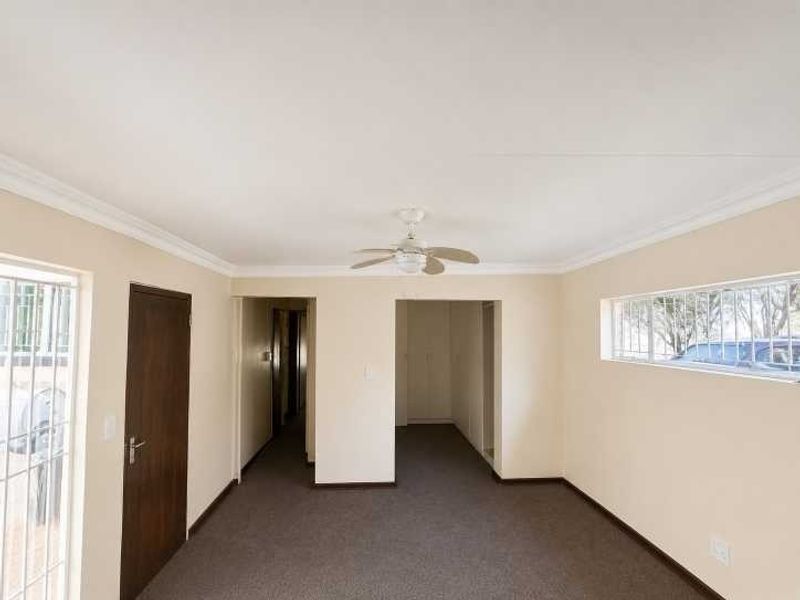 An empty, neutrally painted living area featuring carpeted flooring, a ceiling fan, and security bars on the windows.