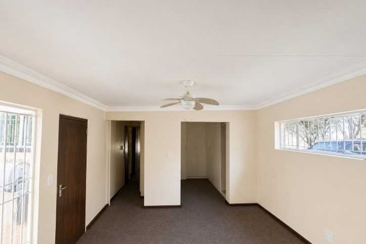 An empty, neutrally painted living area featuring carpeted flooring, a ceiling fan, and security bars on the windows.