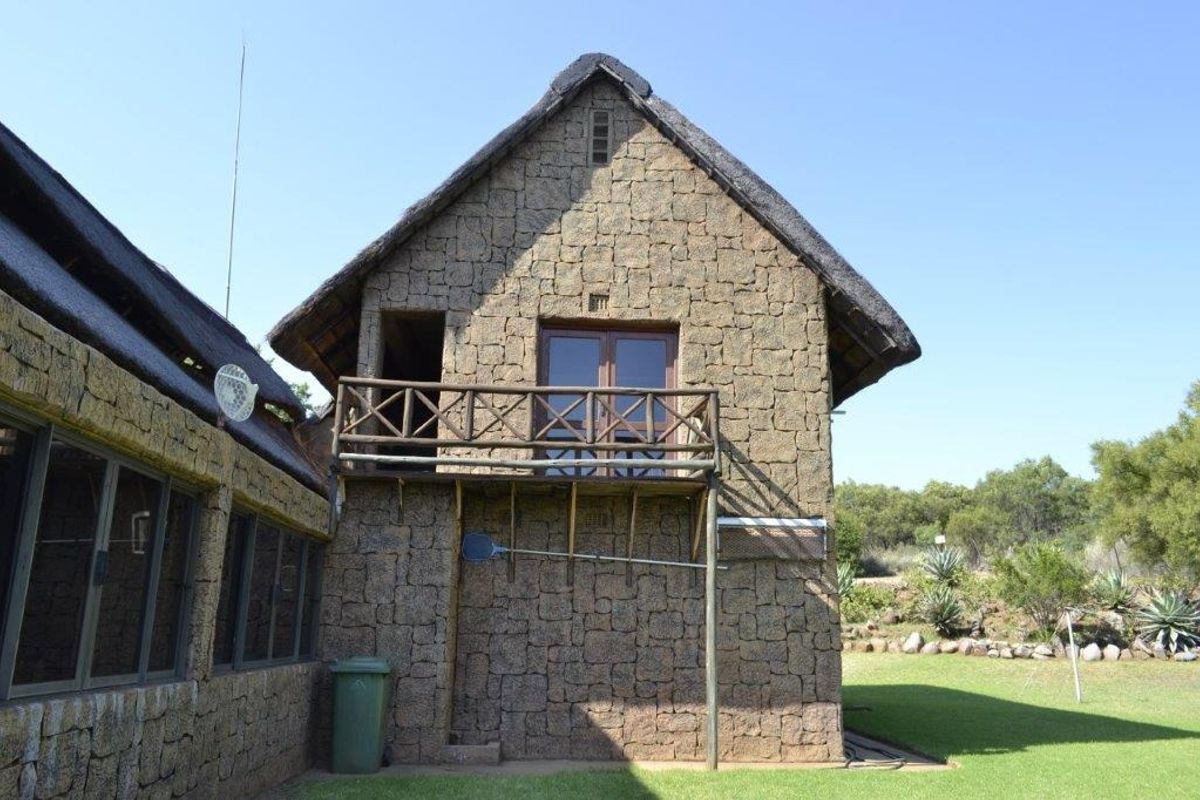 Exterior view of a rustic stone-clad farm building featuring a thatched roof and an elevated wooden balcony overlooking the surrounding garden area.