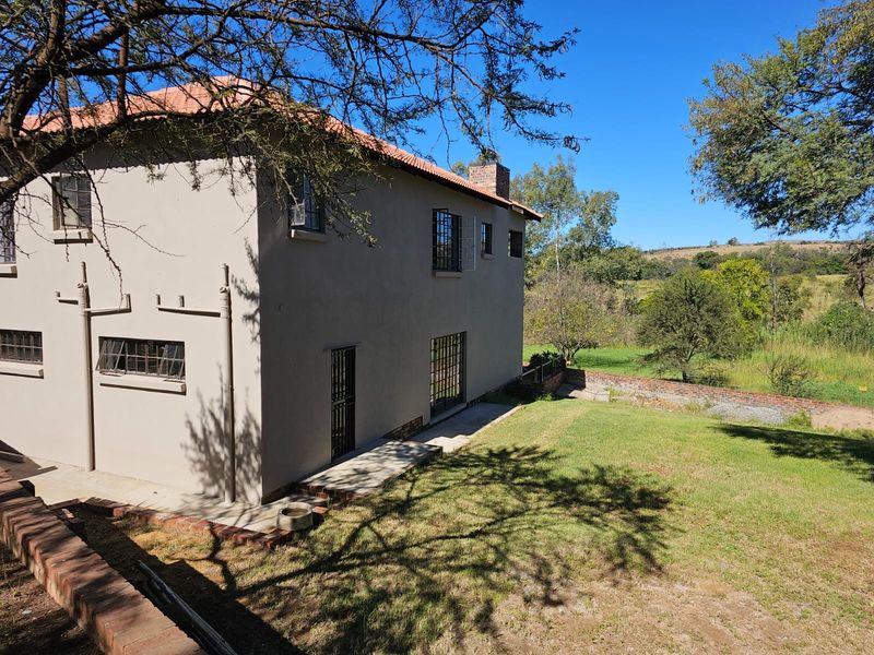 Rear view of the farmhouse featuring a tiled roof, brick chimney, and security bars on windows, overlooking a spacious garden and rural landscape.