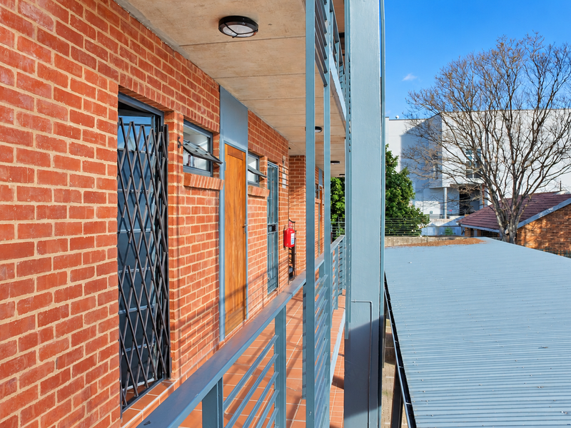 An exterior view of an apartment balcony featuring red brick walls, metal security gates, and a metal balustrade overlooking the complex grounds.