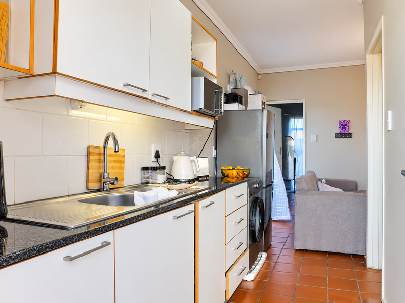 A functional kitchen featuring white cabinetry, granite countertops, a stainless steel sink, and terracotta floor tiles, with an integrated washing machine.